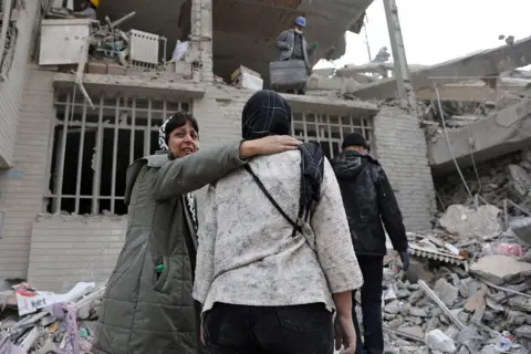 WANA via Reuters A woman holds the shoulder of another woman in front of a collapsed building with rubble all around in Tehran on 12 March.