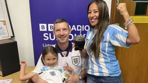 Matthew with a white England shirt sits in a chair in the BBC Radio Suffolk studio with Amelie on his lap. He has grey hair and she has brown hair tied back. Silvana standing next to him is wearing an Argentina blue and white stripe top and blue jeans, and is raising her left fist. She has long dark hair. All are smiling. 