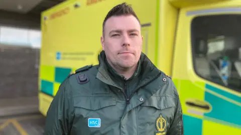 Johnny Hanna, a paramedic, in a green coat in front of a Northern Ireland ambulance. 