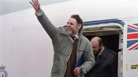 Getty Images SirTerry Waite with medium-length dark hair and beard. He is wearing a green coat over a brown sweater and brown tie. He is waving with his right hand while standing on the top step of passenger stairs attached to an aircraft which has a union flag painted next to the door. There is another man with a beard behind Sir Terry.