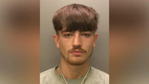 A mugshot of Noah Hetherington. He has brown hair that is longer on the top and shaved at the sides. He has blue eyes and a goatee and is wearing a grey jumper with two necklaces - a golden chain and one with white beads. He is looking at the camera with a serious expression. 