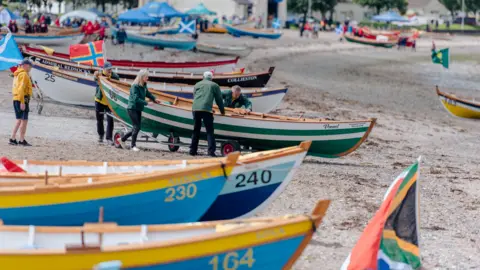 A number of rowing boats are lined along the shore. They are all of different bright colours. A group of people are by the side of one of the boats.