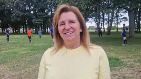 Naj Modak Sue wearing a light yellow top, standing on a grassy park field while football training activities take place behind her among trees and training cones.