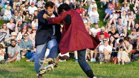 Two men competing in the shin-kicking event at the Cotswold Olimpik Games. They are holding onto each other's shoulders as they kick at their opponent's padded shins.
