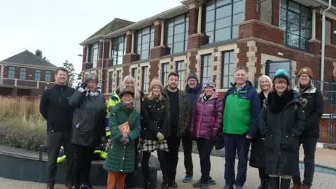 Westmorland and Furness Council A group of 12 people standing in front of a large brick building with large windows. They are all looking into the camera and smiling and wearing various coloured coats and hats.