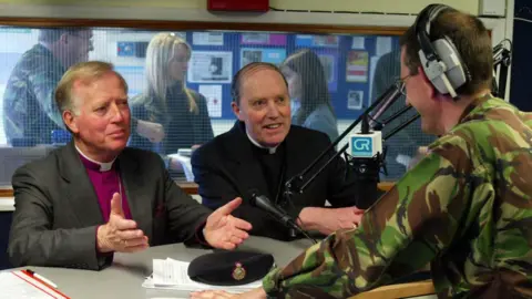 PA Media Inside a radio studio, a person in a camouflage military uniform is leaning over a control panel with audio equipment, engaging in conversation with two men seated at a table. They are the Bishop of Chelmsford, who is wearing a clerical outfit with a purple shirt and a white collar, and Bishop McMahon, who is dressed in black clerical attire.