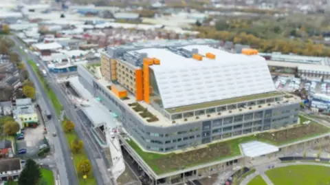 Sandwell and West Birmingham NHS Trust Aerial view of a very large rectangular building, with grey floors at the bottom, and a white roof with orange elements to the side and top.