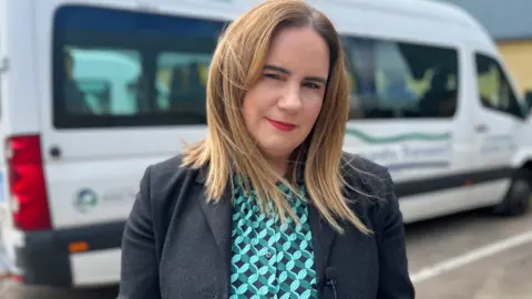 Noeleen Lynch looking directly at the camera, with one of her organisation's buses behind her. She has long blonde hair and is wearing a blazer