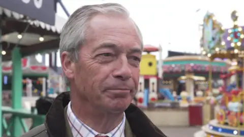 Nigel Farage, who is wearing a green wax jacket and shirt, smiles while stood outside at an amusement park. Rides, including a carousel, can be seen in the background. 