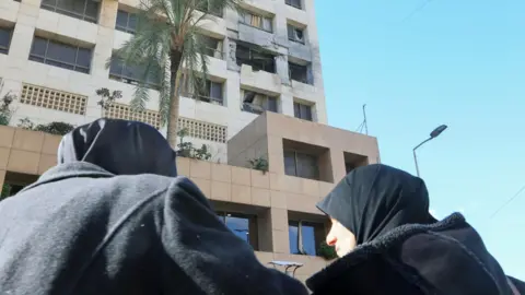 EPA Two women wearing black hijabs stand near the damaged Ramada Plaza hotel following an Israeli strike that the Israeli military says killed four Iranian Revolutionary Guards, in Raouche, Beirut, Lebanon (8 March 2026)