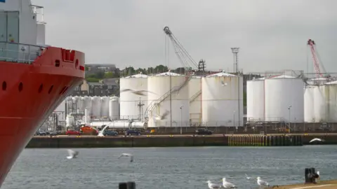 A harbour with a oil plant in the distance and seagulls flying in the foreground by the hull of a red boat