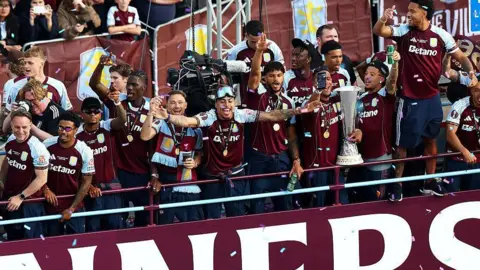 Aston Villa players celebrate with the Europa League trophy on the top deck of a bus as confetti rains down.
