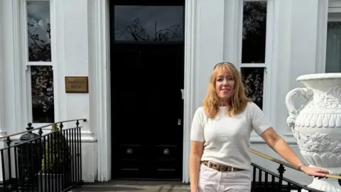 A woman wearing white standing in front of a white georgian house. She has blond hair and is wearing a belt and hand on the black railings. The house is large and has old georgian windows. 