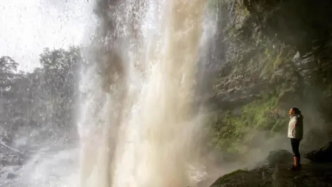 This girl is walking A woman is standing behind a large waterfall and watching it fall from above