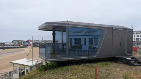 A large grey cabin with a balcony overlooks a beach that has a white lifeguard station. 