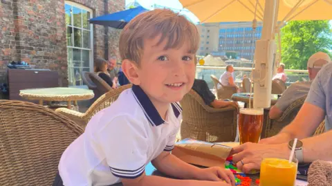 PA Media Young boy in white polo t-shirt smiles into camera as he leans over the table with an orange juice in front of him.