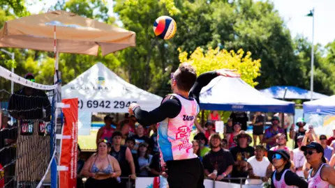 Kaktus Scot A beach volleyball player jumping into the air, arm outstretched to hit the ball over the net as onlookers watch from the sidelines