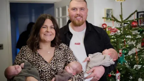 ITV News Stacey and Grant standing together, smiling to camera, with mum holding two triplets, and dad the other one; all are fast asleep. There's a Christmas tree in the room in the background  