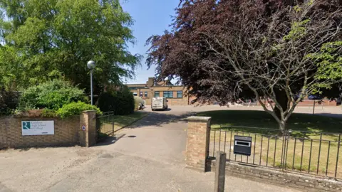 Google Front of school gates showing white sign which reads Reepham High School & College, a driveway, trees and a parked white van