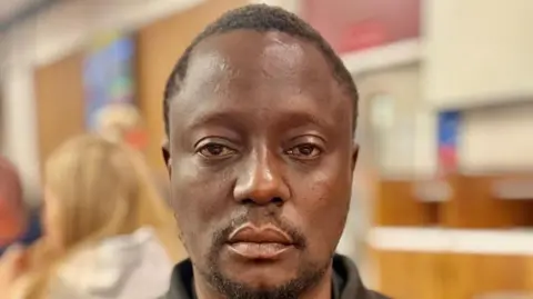 A headshot of Godwin Kayangwe, looking directly into the camera with a serious expression on his face. He has short dark hair and a moustache and beard.