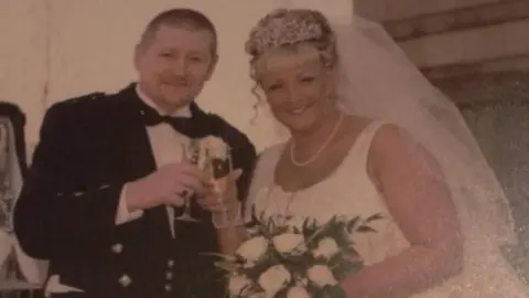A man in traditional Scottish dress including a black bow-tie, waistcoat and kilt, stands next to a woman in a white wedding dress and veil. Both are smiling and are clinking glasses of champagne.
