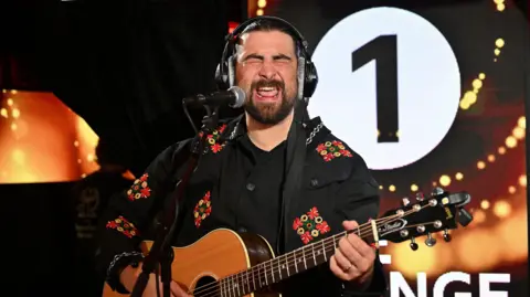Noah Kahan performing in a studio session. playing an acoustic guitar and singing into an microphone. He has dark hair, a beard and is wearing a dark jacket with red, green and yellow markings on it.