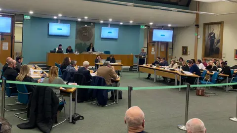 BBC Dozens of people are sat in the council meeting room at rows of desks. They are facing three people who are sitting at a head table at the back of the room. There are screens dotted around the chamber and a painting of a man in military wear on the right-hand side.