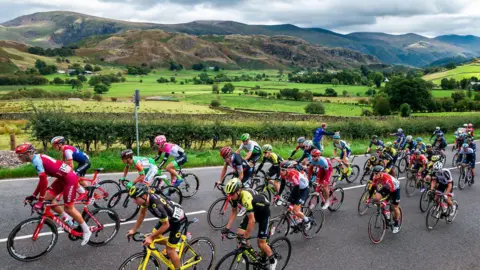 Alex Whitehead/SWpix.com About two dozen riders are cycling up a grey tarmac road with the rolling hills and peaks of the Lake District behind them. The cyclists are in various colours.