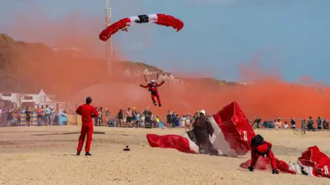 Bold Media Agency Red Devils parachutists landing on Bournemouth beach with spectators watching from behind barriers. The parachutists are wearing read overalls and have red and white parachutes and are trailing red smoke behind them