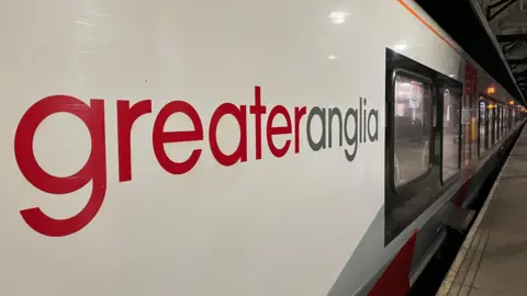 Close up image of a Greater Anglia train - looking along the platform with the company's red and grey logo on the side of the train taking up most of the image.