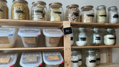 ELLEN KNIGHT/BBC Six wooden shelves can be seen. Four of them have large glass jars on them, with black labels detailing what's inside. In the jars are things like various types of dried pasta and noodles, as well as varieties of flour and yeast. There are two more shelves which have large plastic 'bins', also containing dried pasta. 