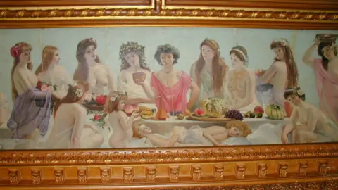 Pauline Hockley A painting shows a group of women having a meal and standing around a table. They are depicted in the classical style, some wearing cloth dresses and some with exposed bodies.