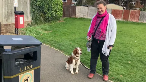 BBC Paula is seen wearing a pink scarf and light cardigan stands on a park path holding her dog’s lead beside a litter bin. The brown-and-white spaniel sits at her feet.