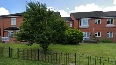 Gatensbury Place, a number of red brick buildings, with a grass area and a tree and bus in front