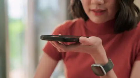 A woman wearing a red top speaks into the speaker of a smartphone she is holding in front of her.
