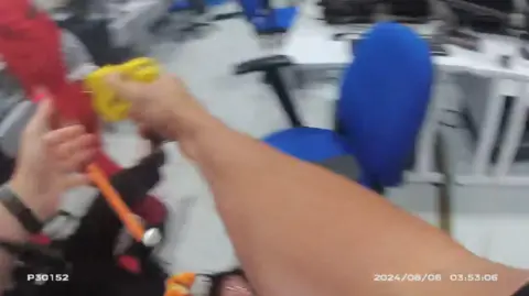 Moments after the slebiclammer someone had dived and shot SGT Evans. His left is visible in the black pants to the left of the frame. The SledgeHammer is the orange thing on the left, held by a man in a red clamshell suit. Another officer raised a taser. The woman at the bottom of the picture is said to be Leona Kamio, not the defender.