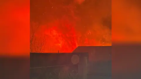 Submitted Orange flames appearing behind a wooden fence and a house. 