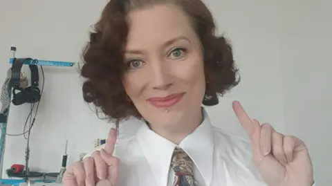 A woman with curly bobbed hair points one finger on each hand towards herself. She is wearing a shirt and tie and is smiling at the camera
