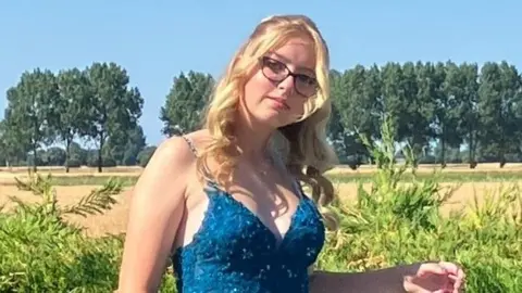 Cambridgeshire Police Eden Bunn looking into the camera on a beautiful sunny day. She has long blonde hair and dark-rimmed glasses and wears a long blue ball gown