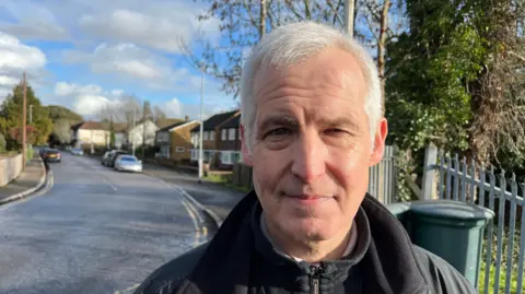 A man stands by a road. There are houses in the background.