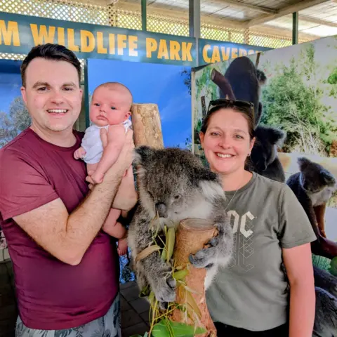 Roddy Graham A smiling couple pose at a wildlife park while holding a baby next to a koala perched on a wooden post eating eucalyptus leaves. The man holds the baby, and the woman stands beside them with sunglasses resting on her head. A sign behind them reads “Wildlife Park,” and large images of koalas are displayed in the background.