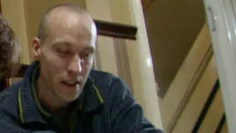 A man with a shaved head is looking down at the floor. He is knelt in the doorway of what appears to be a residential properly. The door frame is painted in cream. He is wearing a blue top.