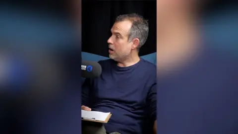 A man with short, grey hair wearing a blue t-shirt sits in front of a microphone
