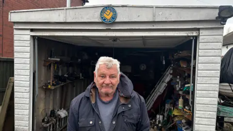 BBC/Jack Conlon A man with white hair stands in front of his open garage. There is a Leeds United ornament with the club's former crest nailed to the top of it. He is stood underneath wearing a grey jumper and a dark grey overcoat.