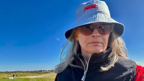 Juanita Adlington is pictured on a golf course. She is wearing a white Jersey Golf bucket hat and a black gilet. She has long grey/blonde hair and big round sunglasses on. 