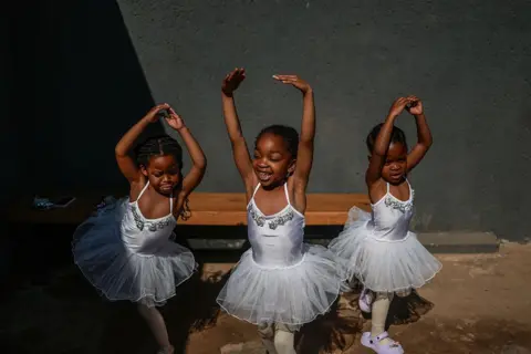 PHILL MAGAKOE / AFP / GETTY IMAGES Three girls wearing white leotards and tutus stretch their arms out.