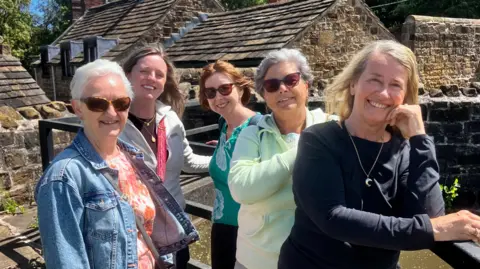 Five women stand along a barrier with a canal behind them. They are all dressed casually in jumpers and smile at the camera.