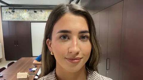 A young woman, Rhian McCrory. She is wearing a plaid top and had brunette hair. Behind her are cupboards and a table.