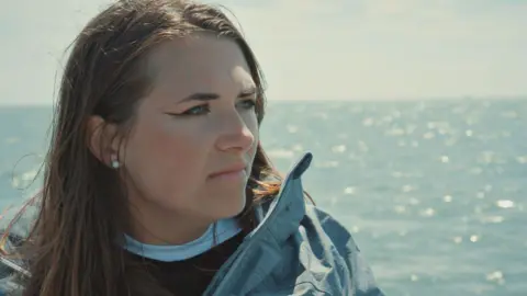 Minnow Chloe during filming Go Back To Where You Came From. Chloe has long brown hair worn loose, pearl earrings and wears a blue waterproof over a dark jumper. She's pictured in front of the sea which is glittering in the sun. 