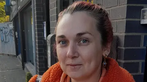 BBC Jamie-Lee Peden wearing an orange coat with brown hair swept back from her head